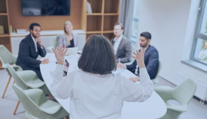 A líder da equipe de vendas está em uma sala de reunião em frente a equipe dando um exemplo de como motivar uma equipe. A equipe é composta por três homens e uma mulher. 