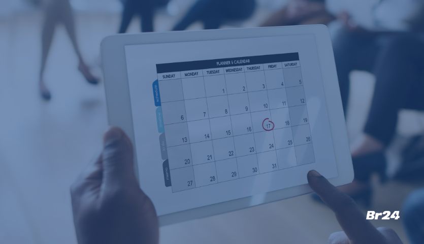 Funcionário olhando calendário corporativo no tablet em uma reunião com outros profissionais no fundo da imagem.
