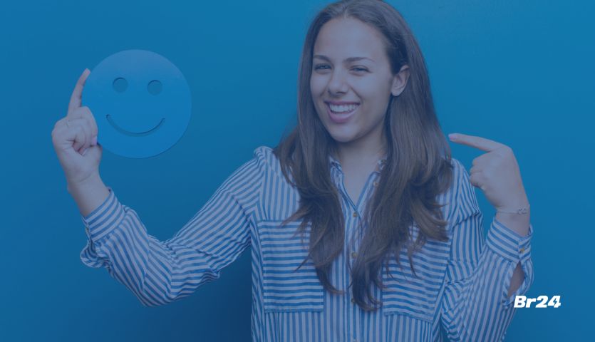 Mulher branca sorrindo apontando para um emoticon feliz em sua mão, representando o customer effort score.