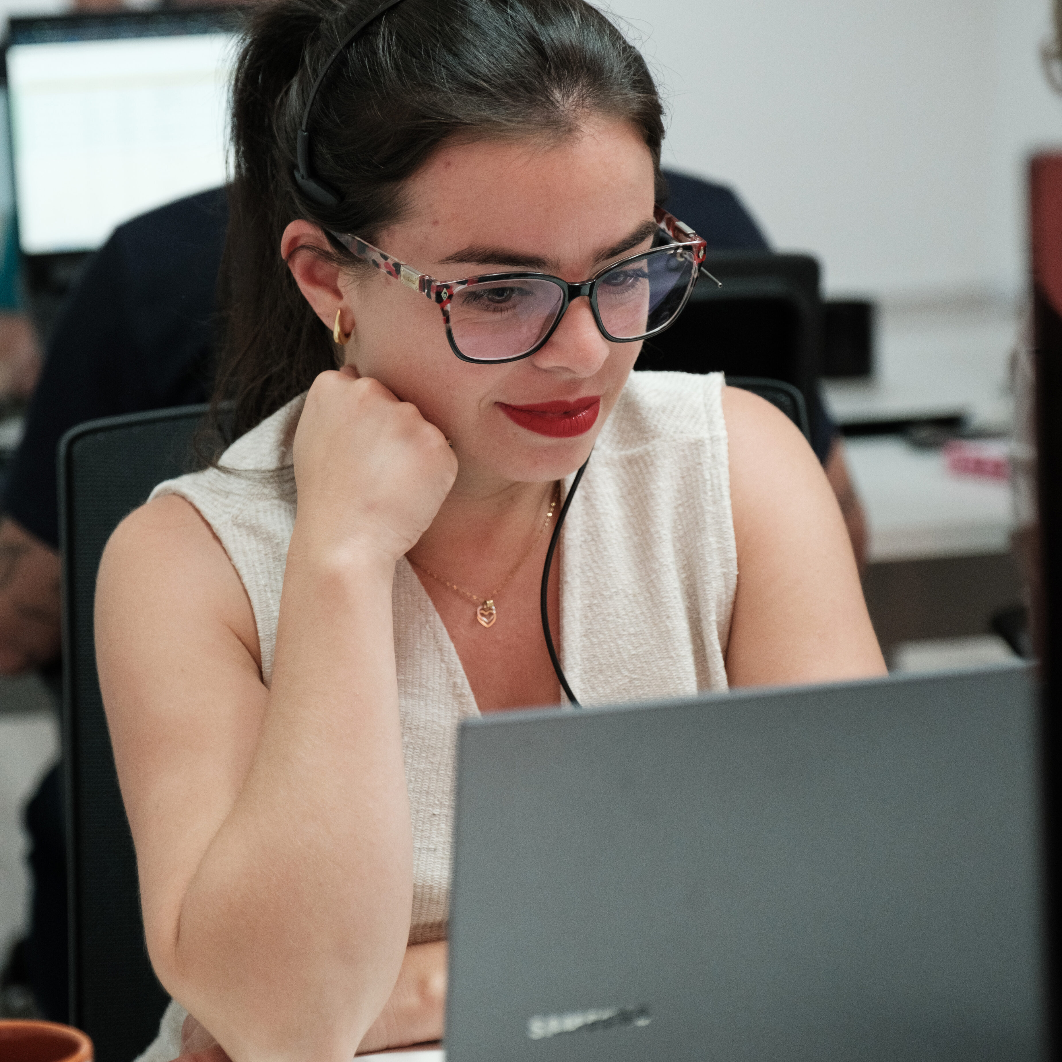 Semana Galáctica Br24. Mulher jovem usando óculos e fones de ouvido, concentrada em frente a um laptop no escritório da Br24. Foco em trabalho e produtividade.