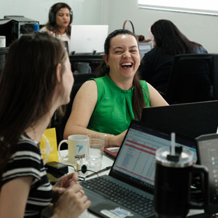 Semana Galáctica Br24. Grupo de shockers da Br24 se divertindo em um ambiente de escritório. Uma mulher sorridente destaca-se com uma blusa verde, rodeada por laptops e canecas, em um clima de descontração e colaboração.