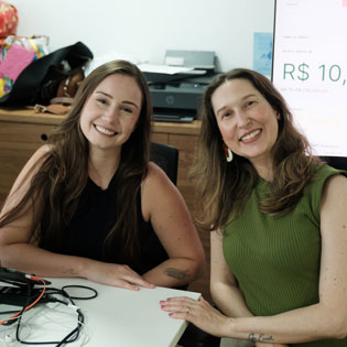 Semana Galáctica Br24. Duas shockers da br24 sorrindo em um ambiente de trabalho, com um computador e materiais ao fundo. Elas parecem felizes em uma interação amistosa.